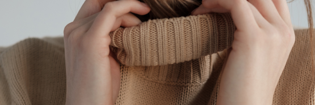 Close up of a woman's neck covered in a tan turtleneck sweater with her hands on the sides of the neck of the sweater