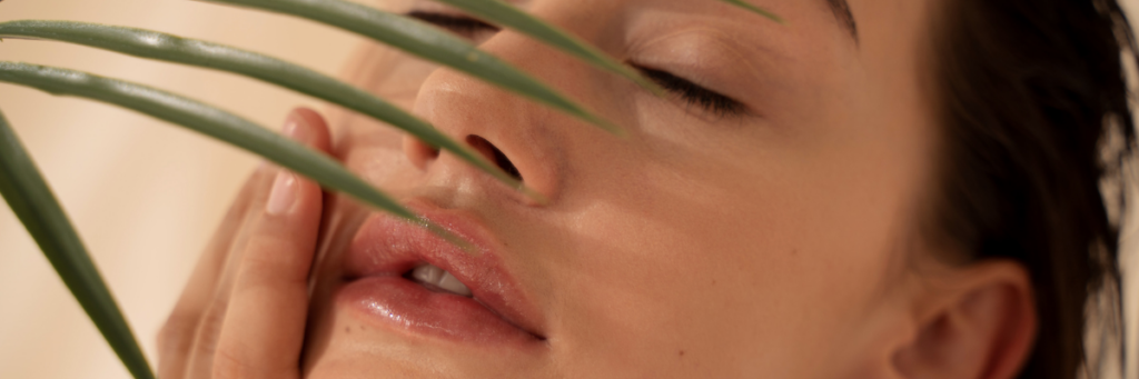 Close up of a woman with dark hair lying with her eyes closed touching the side of her face with her hand with a fern leave casting a shadow on her face