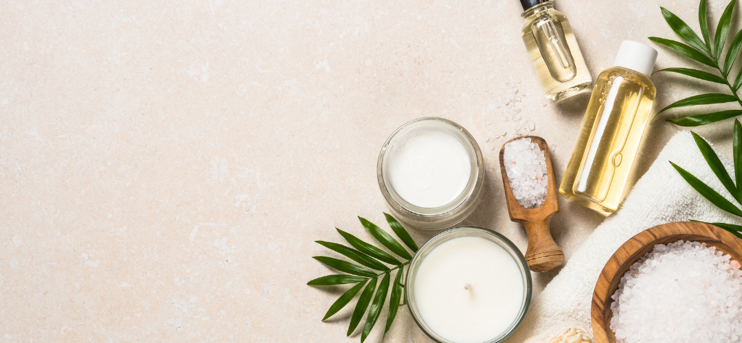Tan background banner with skincare products including a candle, salts, lotion, and oils off to the side with ferns leaves around the products