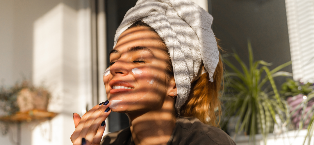 A woman with her hair in a towel and lotion on her face in natural light with window blind shadows on her face
