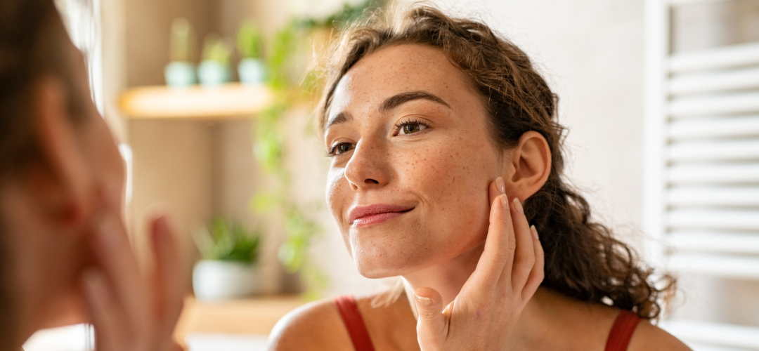 A woman looking in the mirror and smiling while touching her face after a Bela MD treatment