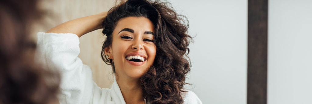 A headshot of a woman looking in the mirror smiling and pulling some of her hair up