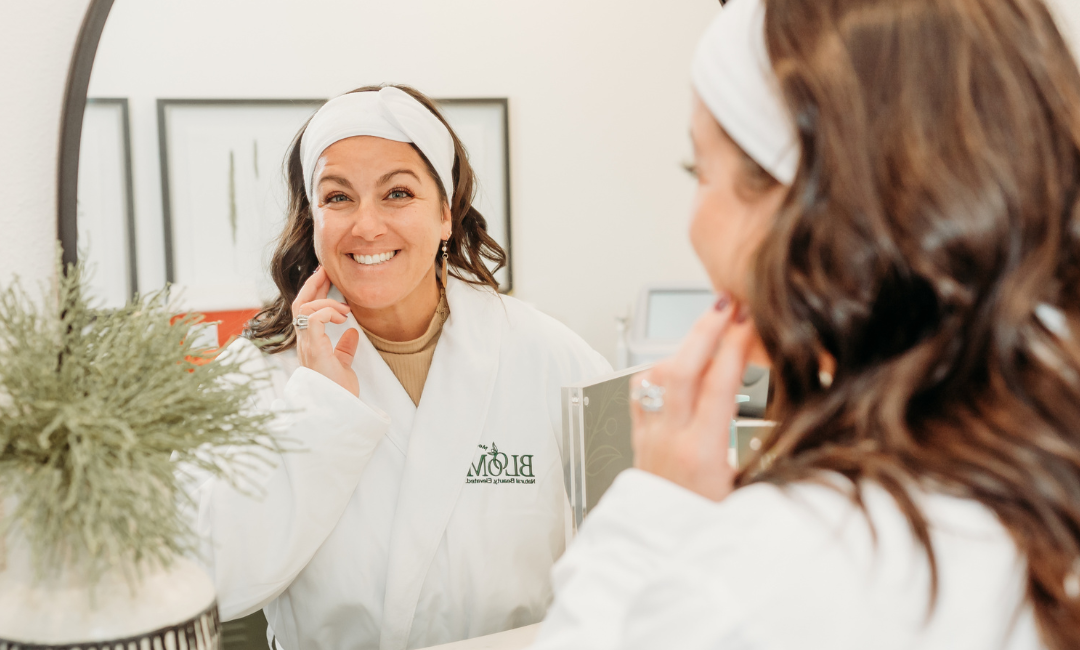 Woman standing with a Bloom MedAesthetics robe on smiling in the mirror at herself